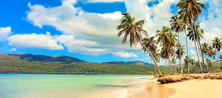 vibrant seascape with turquoise waters gently lapping against a sandy shore under a bright blue sky filled with scattered, fluffy white clouds. In the distance, there are two sailboats with colorful sails, adding a touch of adventure to the serene scene.